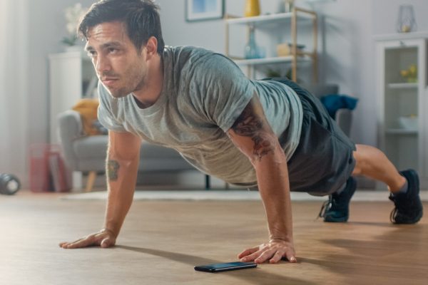 Athletic Fit Man in T-shirt and Shorts is Doing Push Up Exercises While Using a Stopwatch on His Phone. He is Training at Home in His Living Room with Minimalistic Interior.